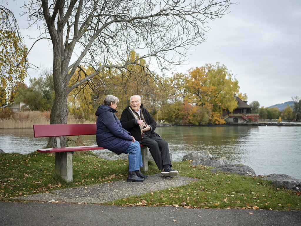 Foto: Älteres Paar im Gespräch auf einer Parkbank am herbstlichen Seeufer